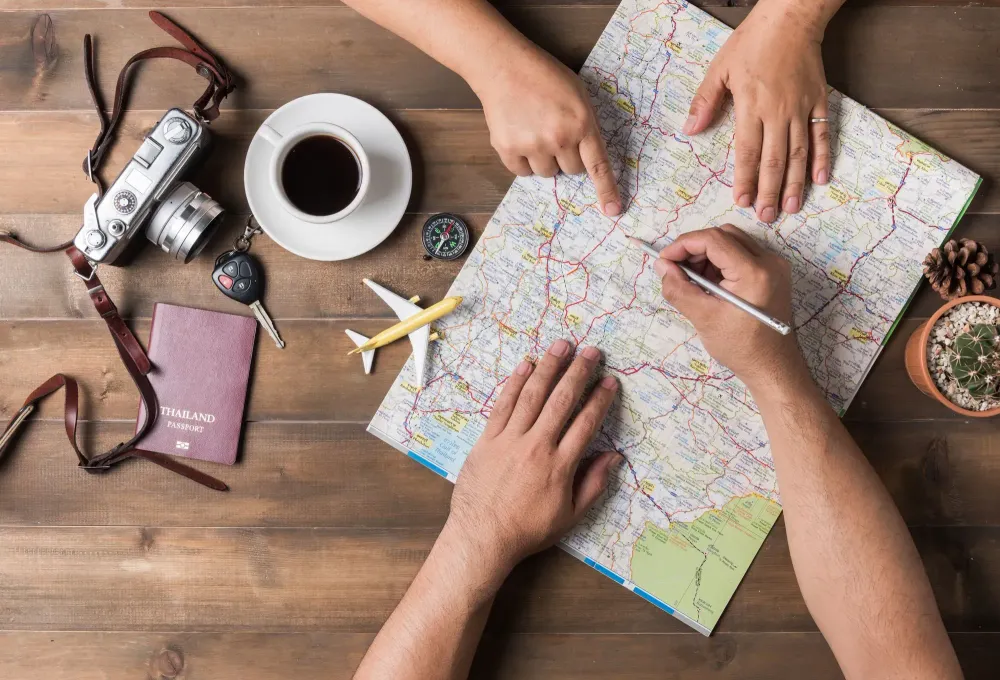 Overhead view of two people planning a vacation on a map surrounded by a cup of coffee, a model plane, a camera, and a passport cover